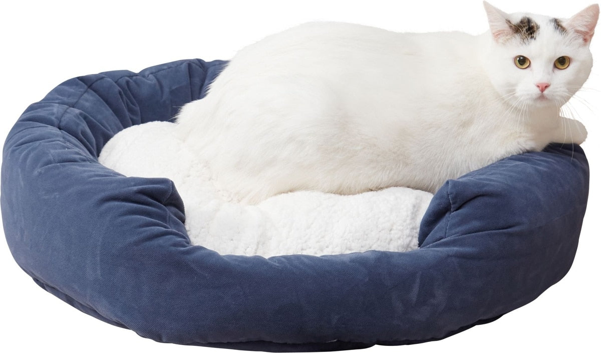 A white cat with black patches lies inside a round, deep pet bed with a navy blue velvet exterior and a white sherpa lining, isolated on a white background.
