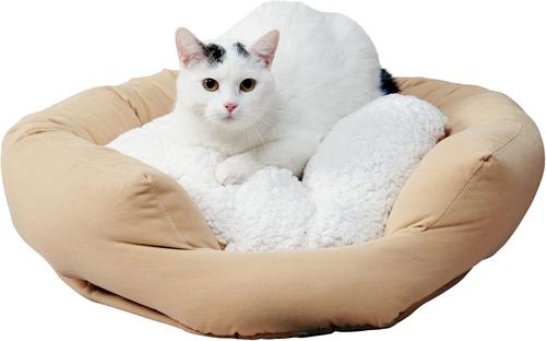A white cat with black patches rests inside a round beige pet bed with a fluffy white sherpa lining, isolated on a white background.