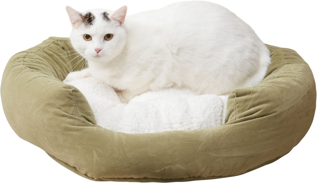 A white cat with black patches lies inside a round, cushioned pet bed with an olive green velvet exterior and a white faux fur lining, set against a plain white background.